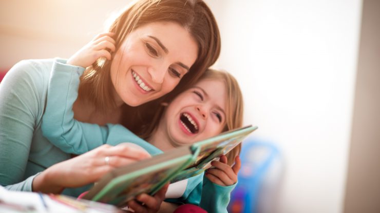 Mother and baby daughter at home reading a book and laughing together.